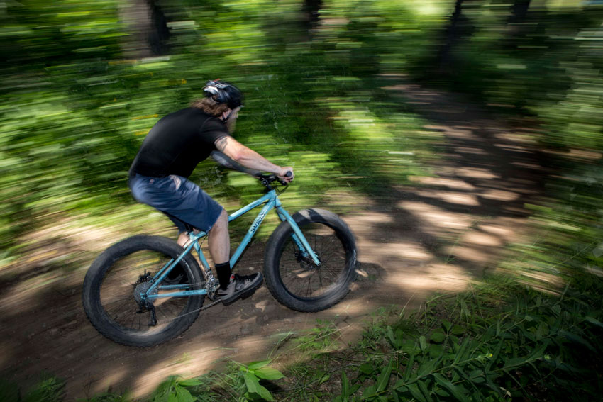 Right side blurred view of a cyclist, on a light blue Surly Wednesday fat bike, riding on a dirt trail in the woods