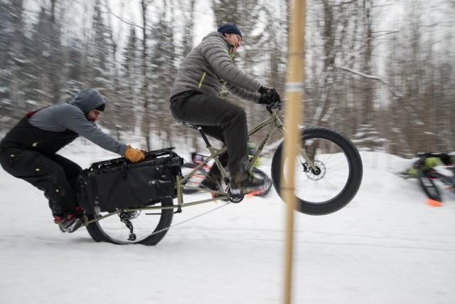 Right side view of a cyclist, riding a Surly Big Fat Dummy bike in the snow, popping a wheelie with a person on the back