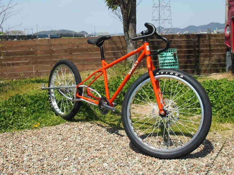 Front, right side view of a modified, red Surly Troll bike, parked on gravel & grass, in front of a tree and wood fence