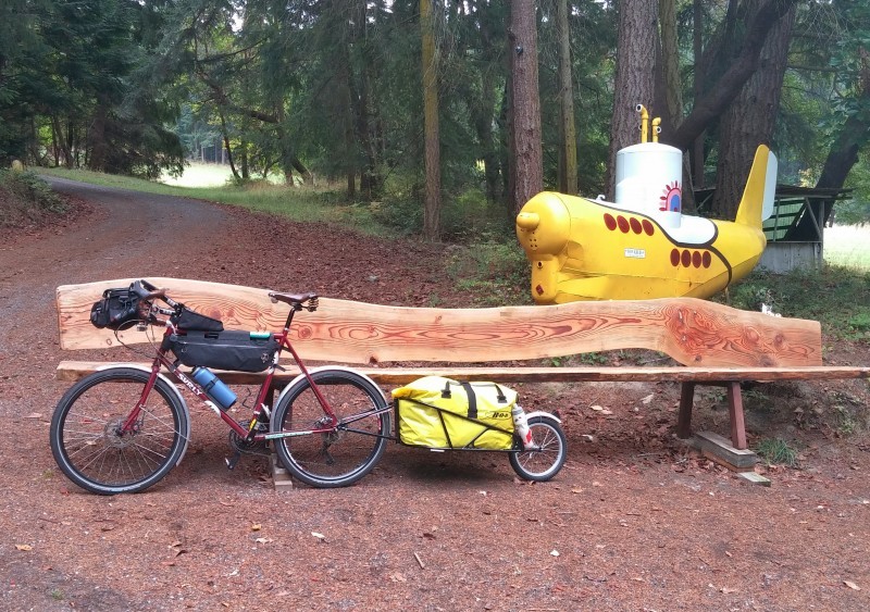 Left side view of a red Surly bike with trailer, leaning on a split log bench, with a yellow submarine statue behind it