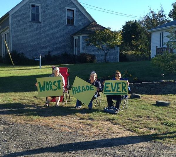 Front view of 3 people sitting in lawn chairs in front of a house, holding up signs that shows, Worst, Parade, Ever