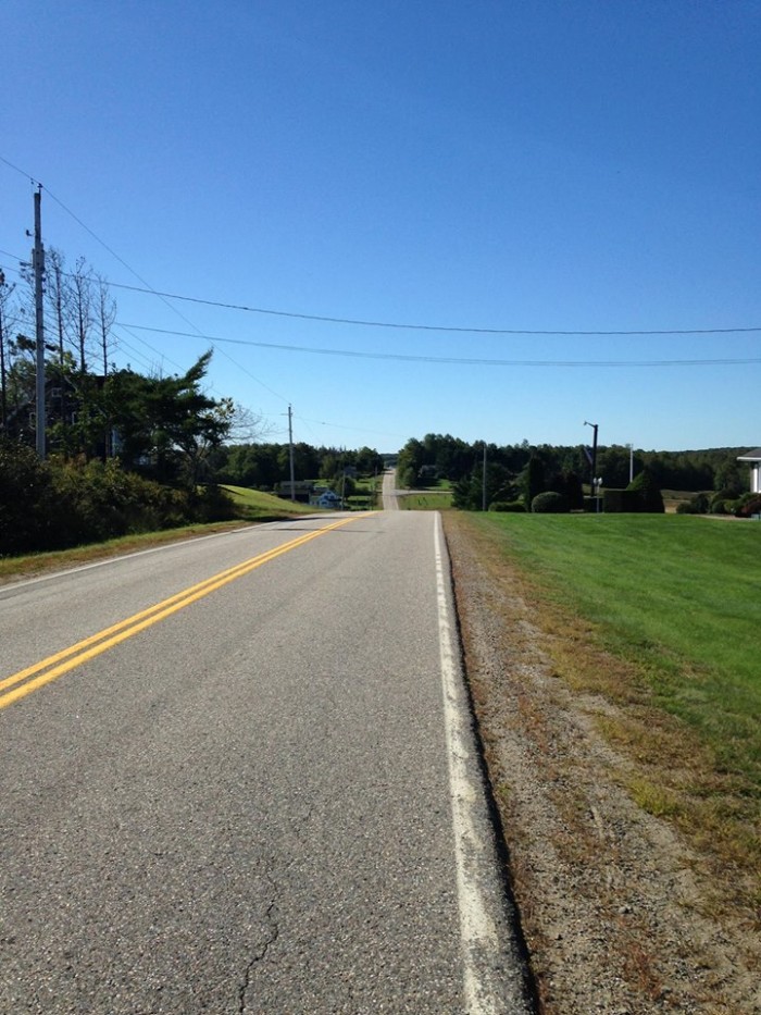 Straight away view of a paved roadway with a grass field to the right, and trees in the background