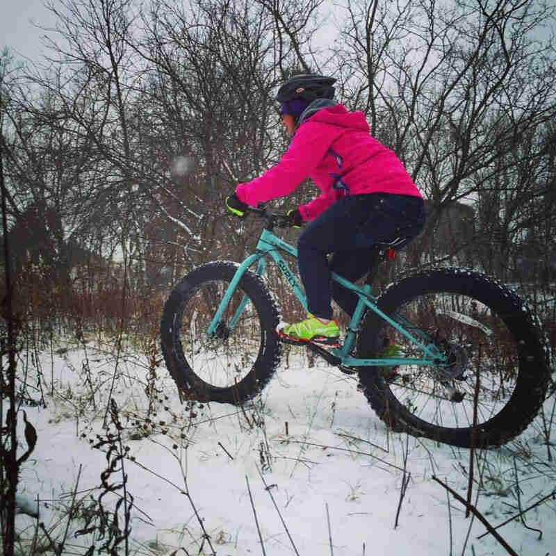 Left side view cyclist riding a turquoise Surly fat bike across a snowy trail in the woods