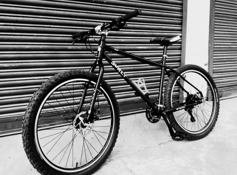 Left side view of a Surly Troll bike, black, on pavement, leaning on a steel garage door - black and white image