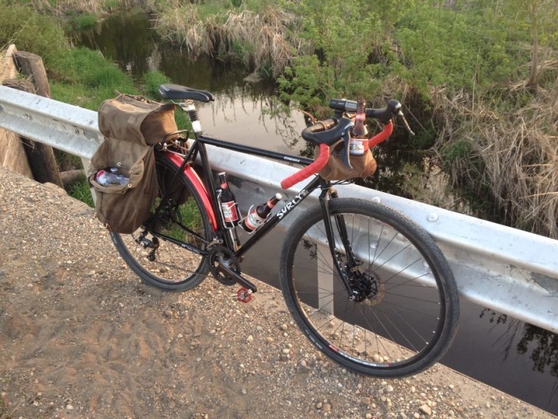 Right side view of a black Surly bike, with rear bags and beer in drink cages, against a rail on a bridge over a stream