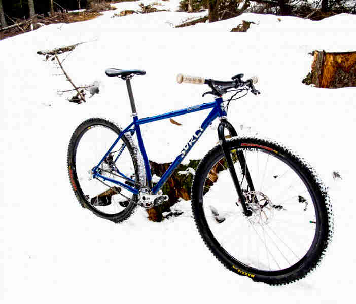 Right side view of a blue Surly bike, parked in a snow covered field, against a tree stump