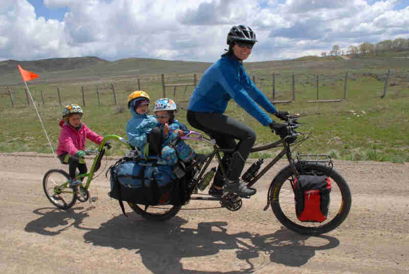 Right side view of a cyclist, riding a Surly Big Dummy bike with 3 children on back, on a gravel road in a rural area