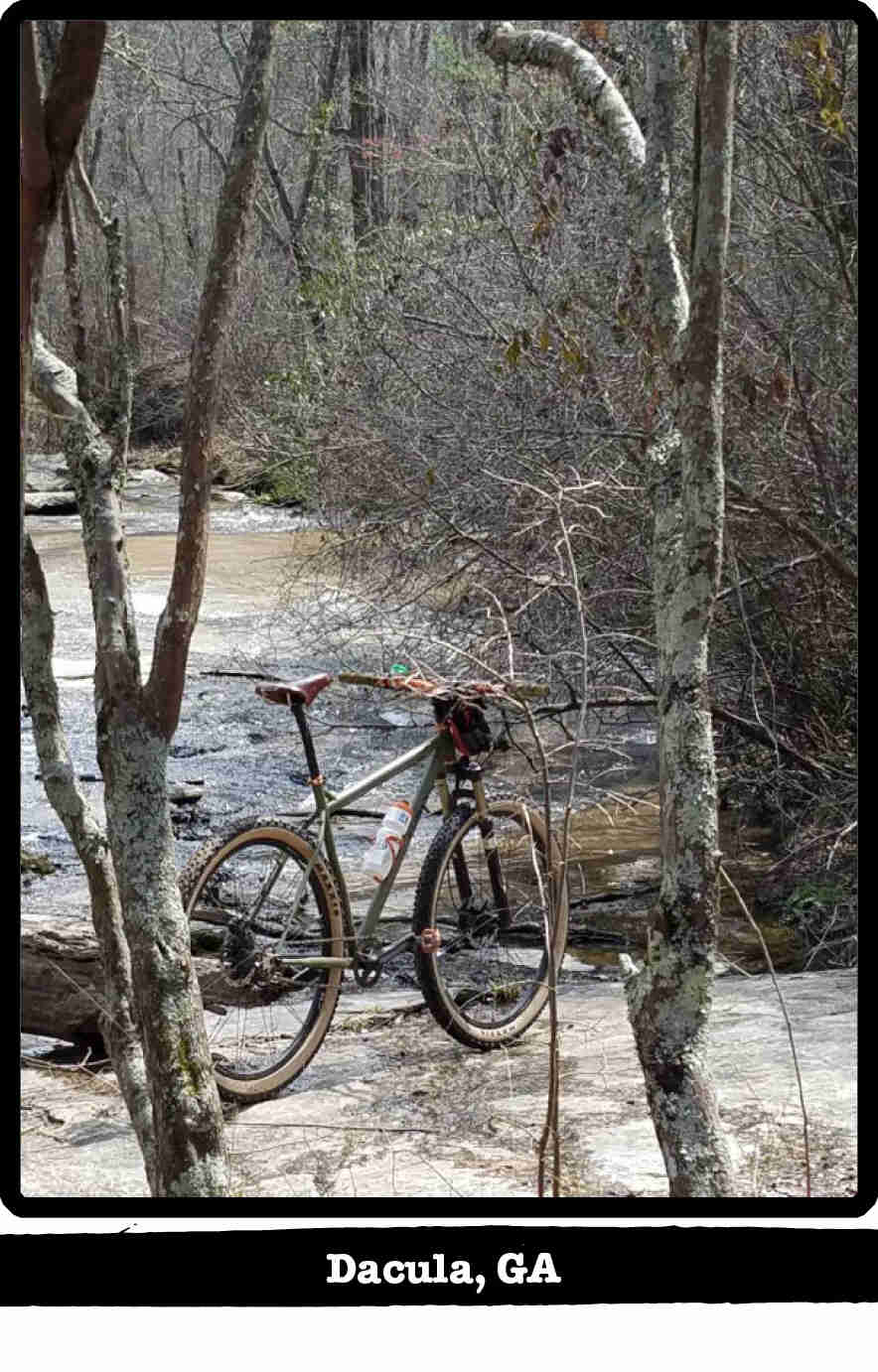 Right rear view of a Surly Karate Monkey bike, olive, at a flat rock on a river bank - Dacula, GA tag below image