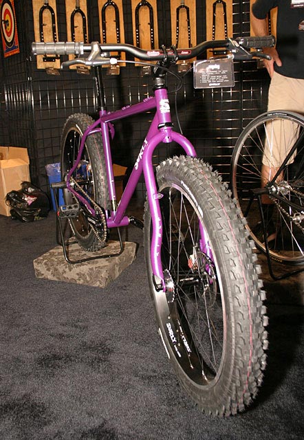 Front, right side view of a purple Surly Pugsley fat bike, in a carpeted room, and bike forks on the wall behind