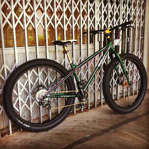 Right side view of a green Surly Krampus bike, on a sidewalk, leaning against a gate in front of a store