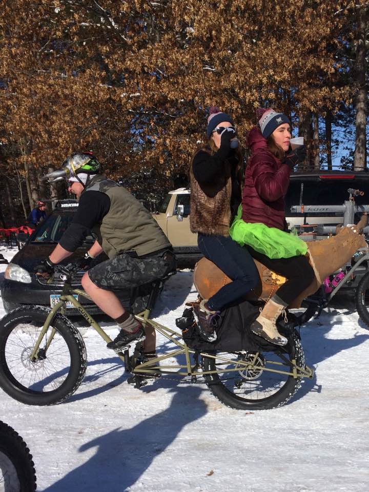 Left side view of a cyclist riding a Surly Big Fat Dummy bike with 2 people sitting on back, on a snowy parking lot