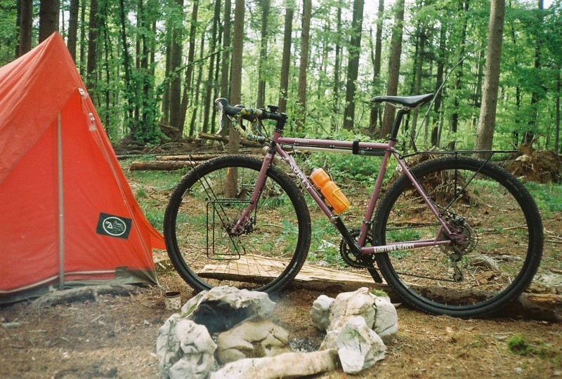 Left side view of a lavender color Surly Straggler bike, next to a red tent on the left, at a campsite in the forest