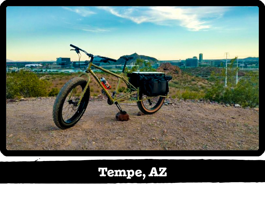 Left side view of a Surly Big Fat Dummy bike, green, on a gravel lot, with city behind - Tempe, AZ tag below image