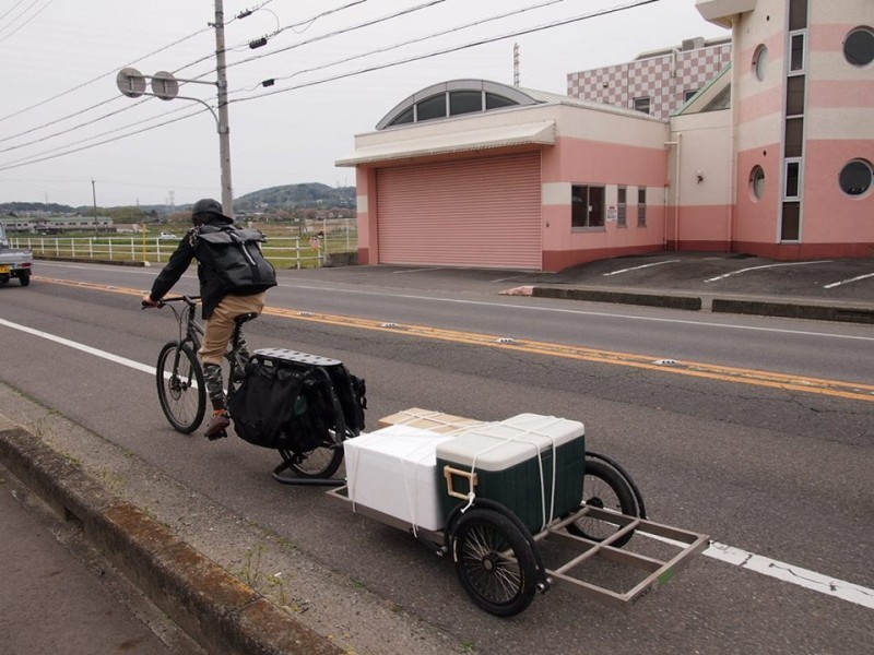 Rear, left side view of a cyclist, riding a Surly Big Dummy bike with a loaded trailer, on the shoulder of a paved road