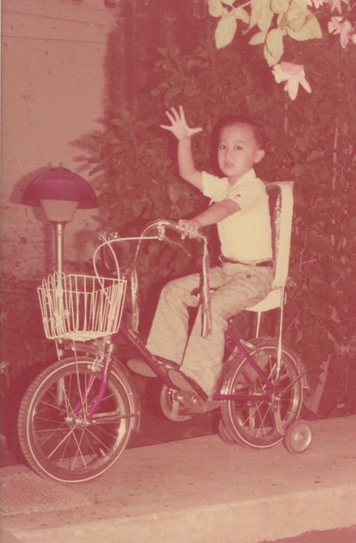 Small child with raised arm on a purple bike with training wheels and front basket on a walkway on the side of a home