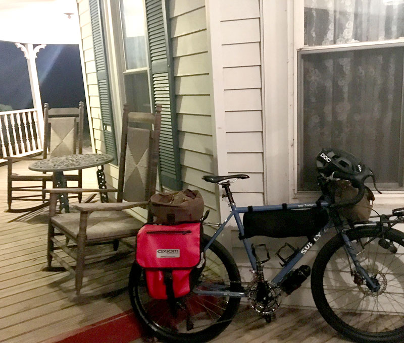 Right profile of a Surly bike, loaded with gear, on a porch against the window of a house with table and chairs behind