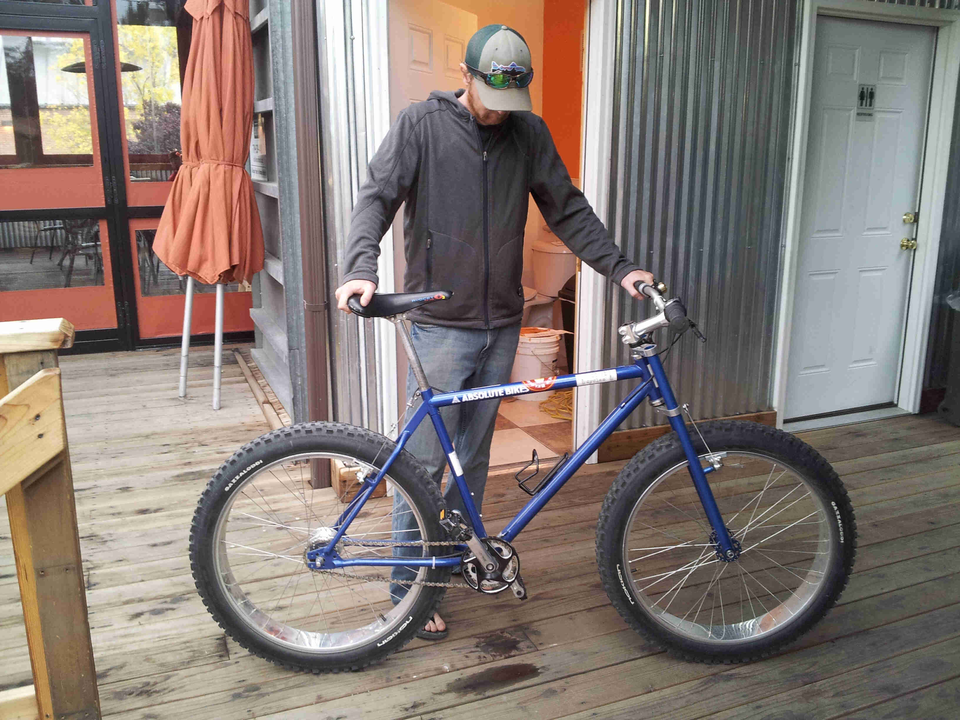Right side view of a blue bike with a cyclist standing behind, on a wood deck patio outside of a building