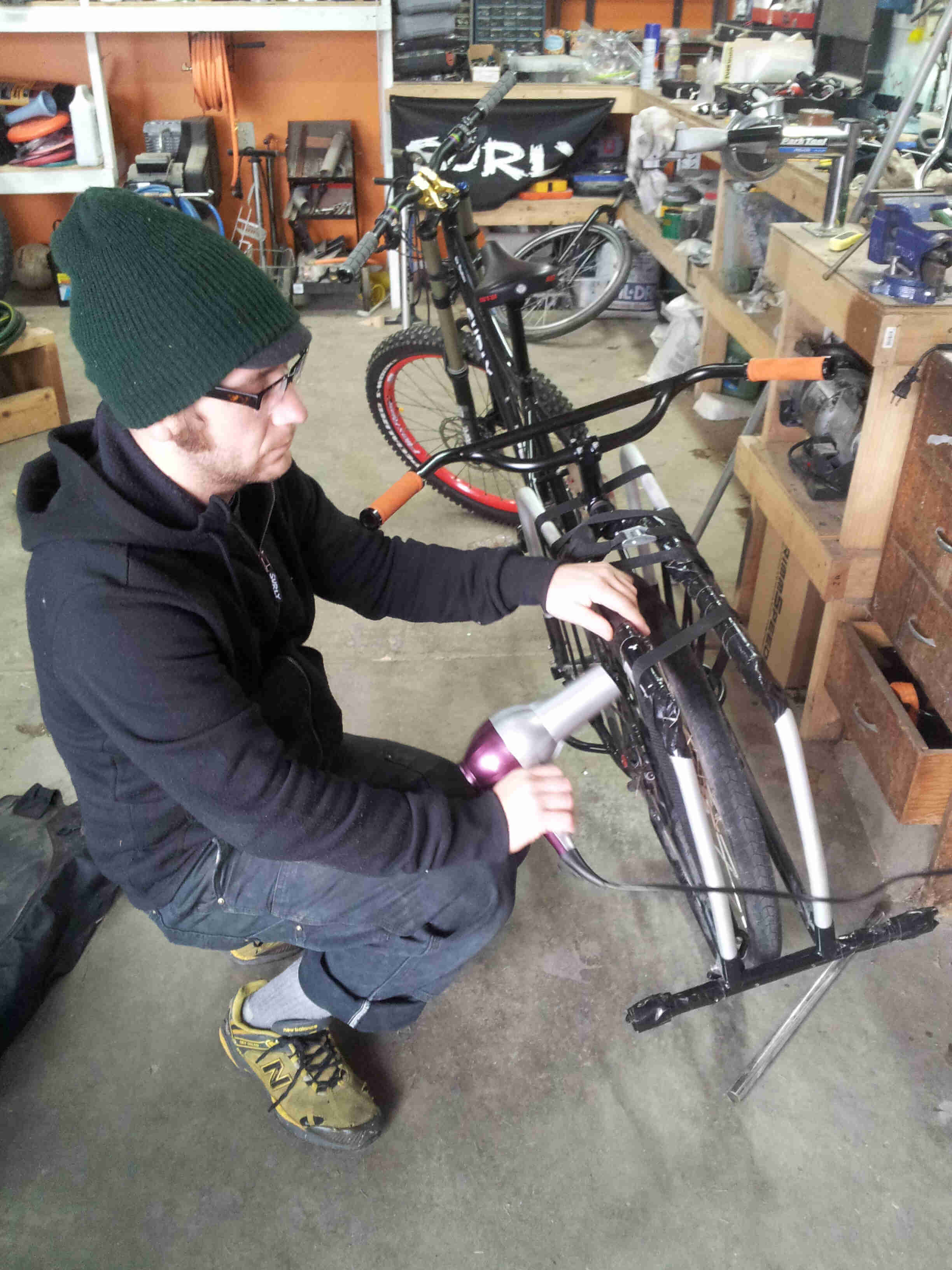 Rear view of a black Surly Big Dummy bike, with a person squatting down on the left side, inside of a workshop