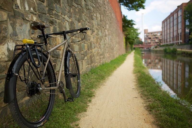 Rear, right side view of a Surly Ogre bike, leaning on a stone wall, next to a dirt trail that's alongside a city river