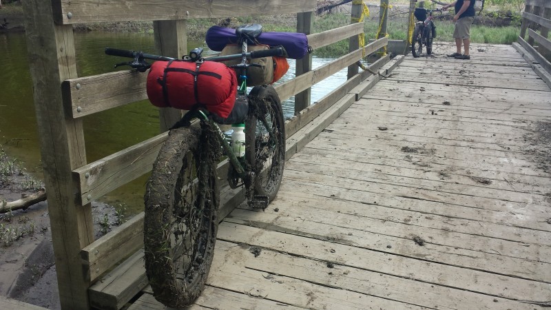 Front, left side view of a green Surly bike with muddy tires and gear, against a rail, on a wood bridge above a stream