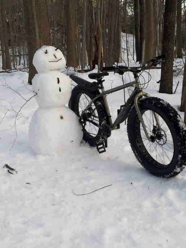 Front, right side view of a Surly fat bike, parked in the snow against a snowman, in the woods