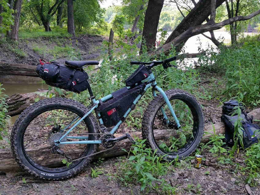 Right side view of a light blue Surly fat bike with gear packs, parked in the woods 