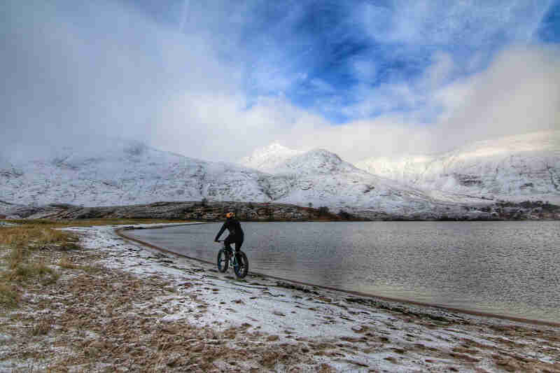 Rear view of a cyclist riding a fat bike along a frozen shore of a lake, with snow covered mountains in the background