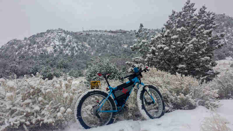 Right side view of a blue Surly fat bike, parked in snow, next to a grass field, with mountains in the background