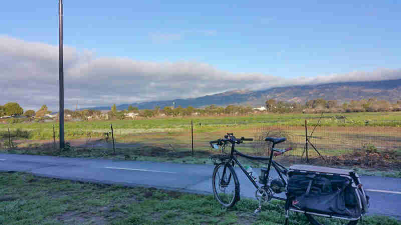 Left side view of a black Surly Big Dummy bike, parked in grass along a trail, with mountains in the background