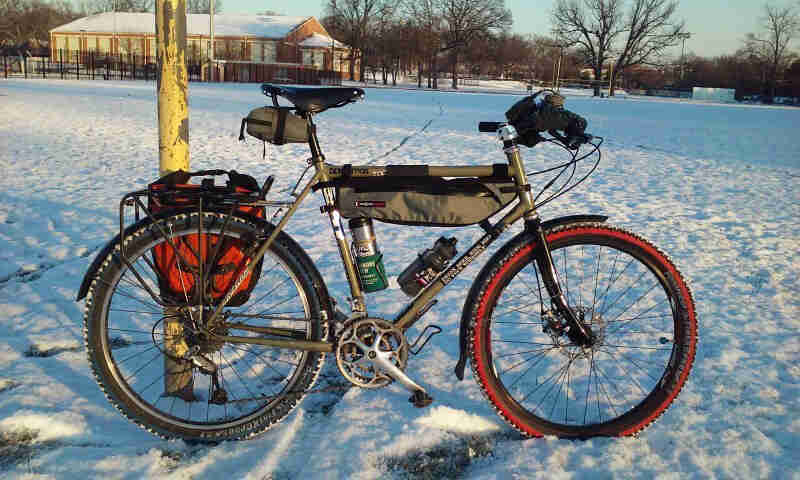 Right profile view of a Surly bike with gear, in a snowy field, with a building an tree in the background