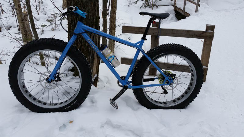Left side view of a blue Surly fat bike, parked in the snow, leaning on a tree