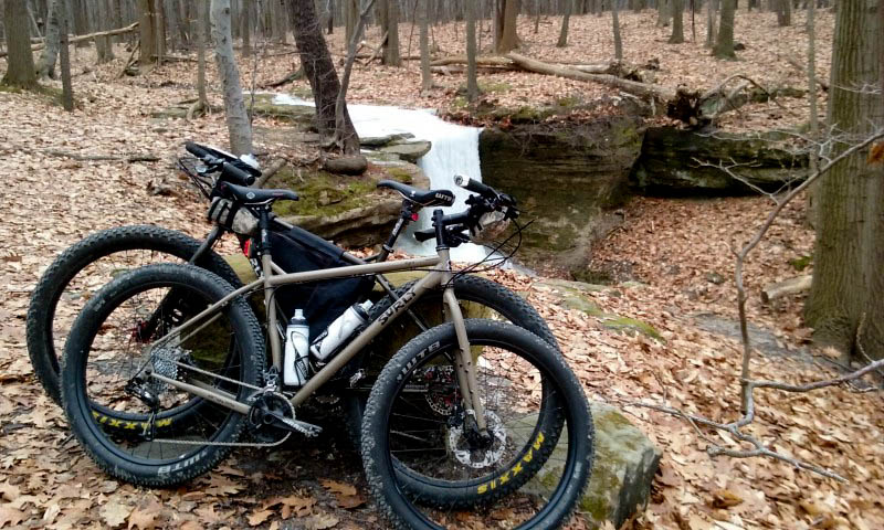 Side view of 2 Surly bikes leaning against each other, on a large rock in the woods, with a stream in the background