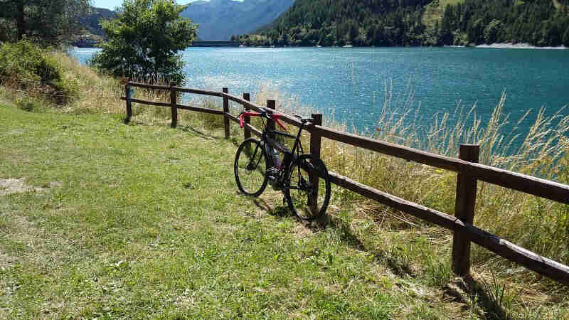 Rear view of a bike parked in a grass field, along a fence, with a lake and mountains on the right, in the background