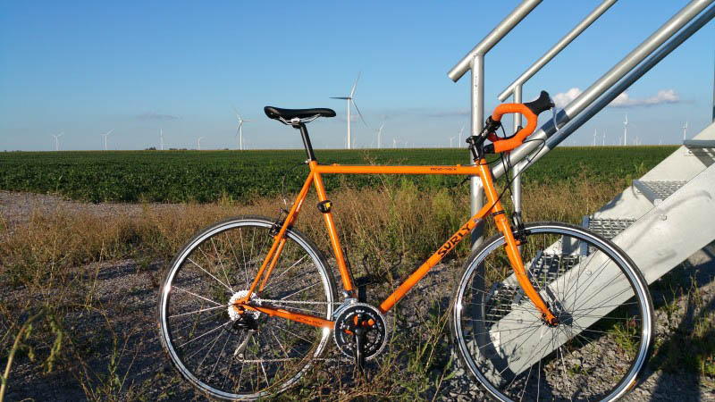 Right profile of an orange Surly bike in front of a set of stairs, and a field with windmills in the background