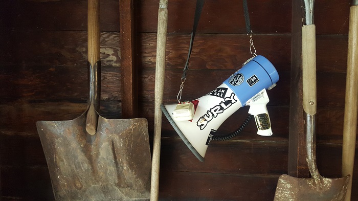 A blue and white megaphone with a Surly sticker on it, hanging down in front of a wood barn wall with shovels on it