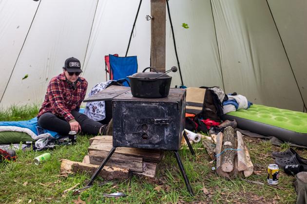 Person wearing a flannel shirt sitting on a sleeping bag next to a wood stove in a tent