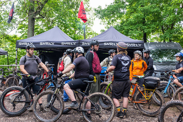 A group of cyclists with their bikes standing in front of two black Surly tents in the woods