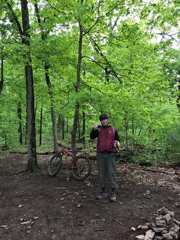 Cyclist standing on dirt with trees behind, holds up two cans
