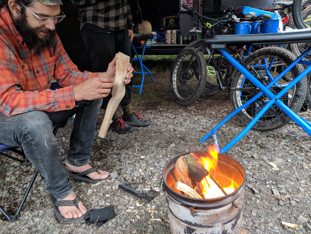 Person sitting in a camp chair holds kindling while looking at a fire in a steel bucket