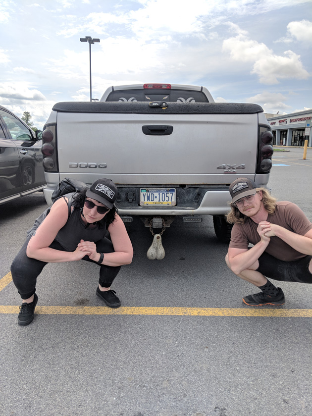 Two peoples dressed in Wayne's World attire squatting down a parking lot behind a truck