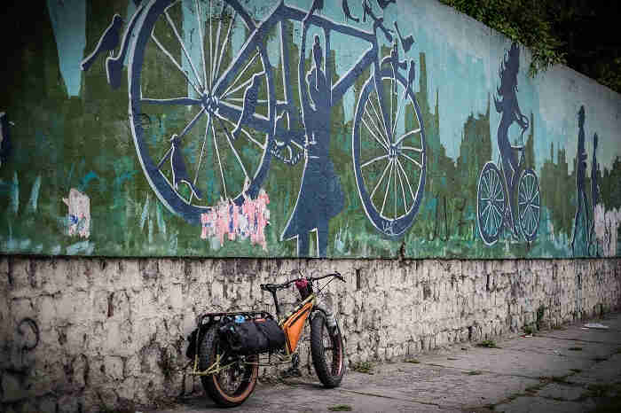 Rear right side view of a Surly Big Fat Dummy bike on a sidewalk, against a wall with a mural