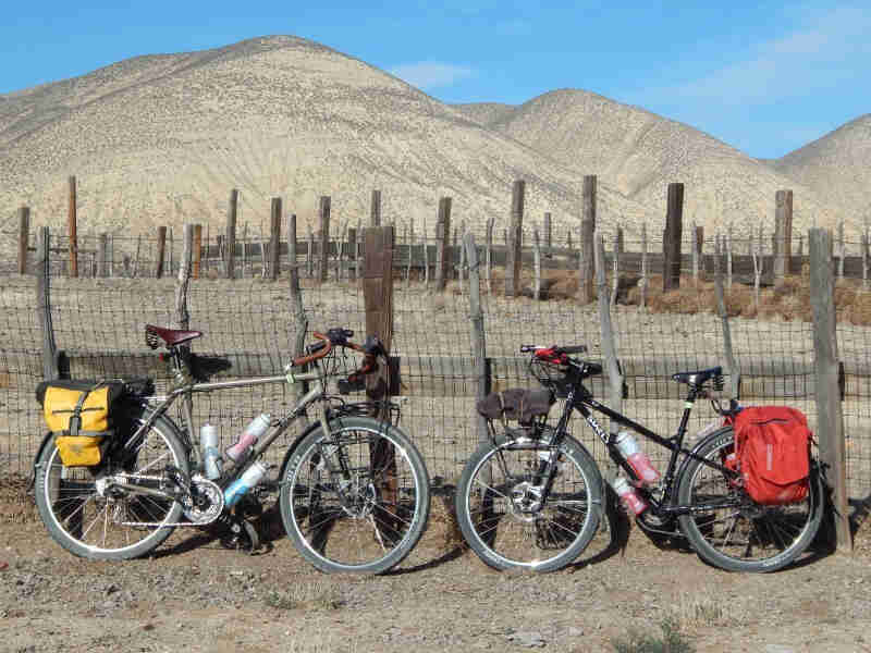 Right side view of 2 Surly bikes, facing each other, on the outside of a livestock pen, with desert mountains behind it
