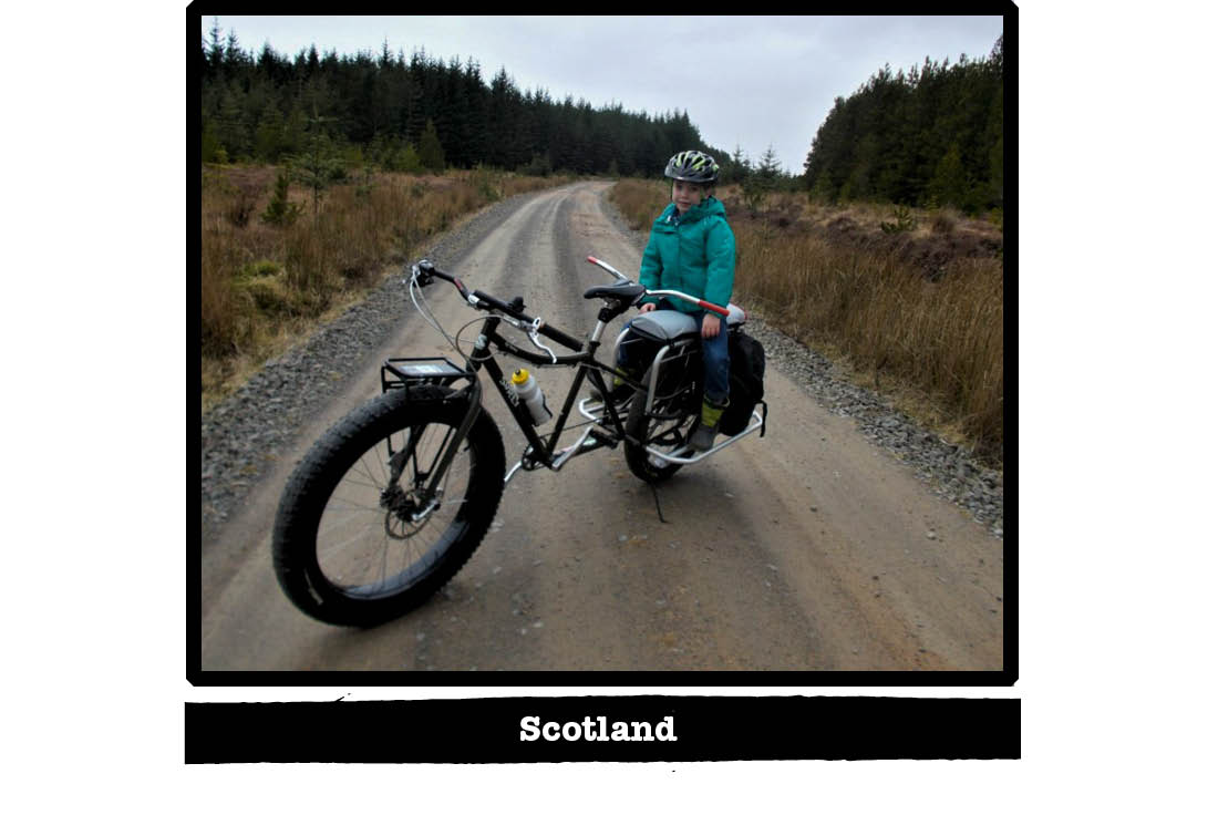 Front left view of a Surly Big Fat Dummy bike with a child on bike, on a gravel road - Scotland tag below image