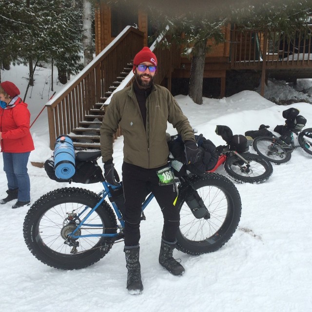 Front view of a cyclist, with their fat bike behind them, standing in snow with a cabin in the background