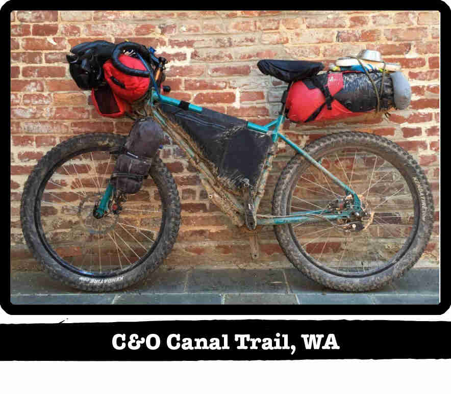 Left side view of a muddy Surly bike with gear, leaning against a red brick wall - C&O Canal Trail, WA tag below image