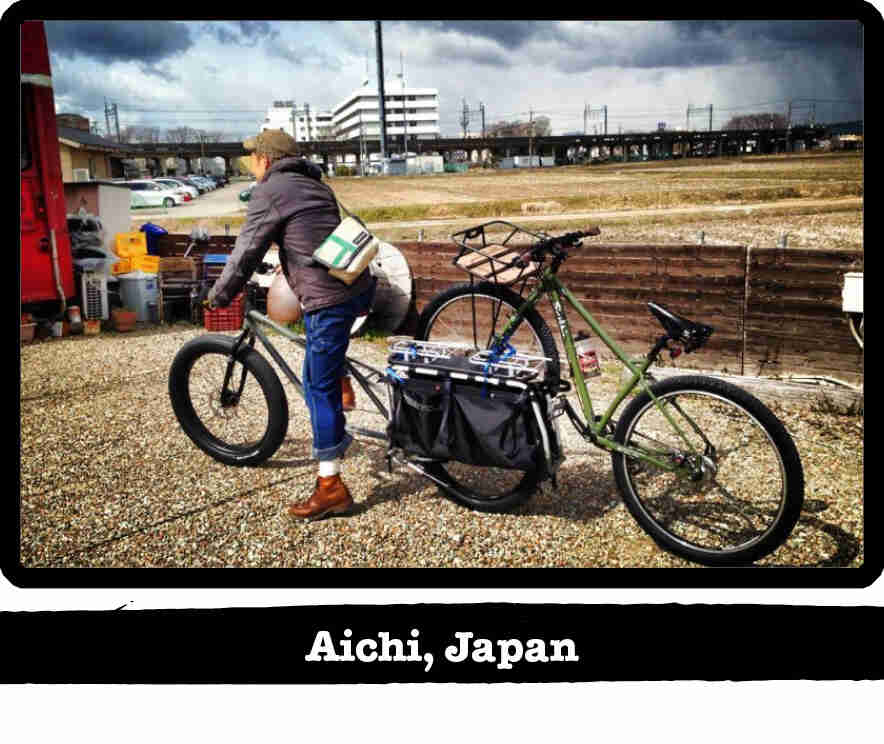 Cyclist on a Surly Big Fat Dummy with another bike on back, with city in the background - Aichi, Japan tag below image