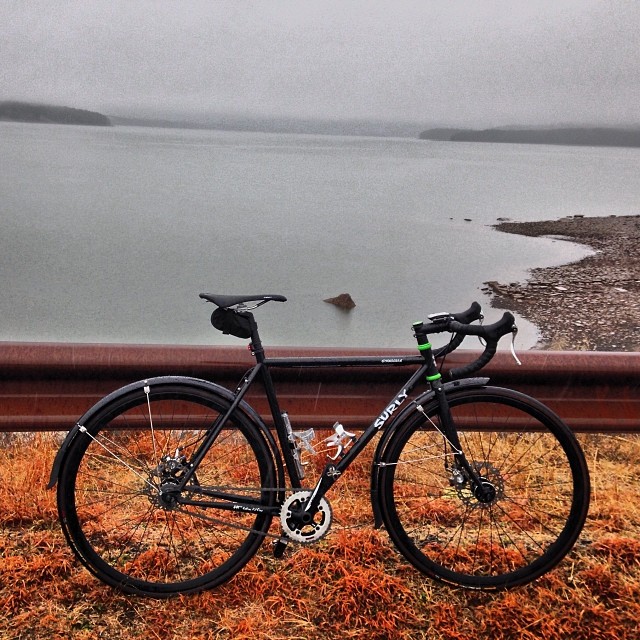 Right side view of a black Surly bike with fenders, leaning on a steel, road guardrail, with a foggy lake behind it