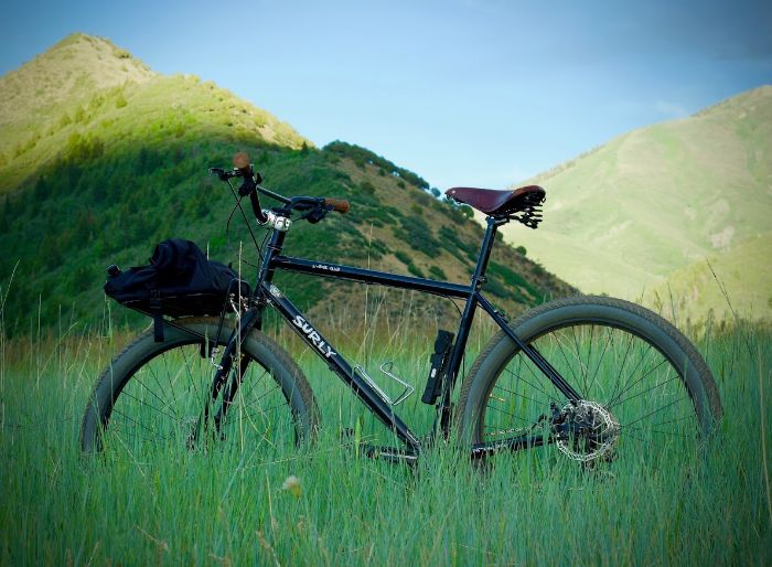 Left side view of a black Surly Bridge Club bike, standing in a field of tall green grass, with large hills behind it