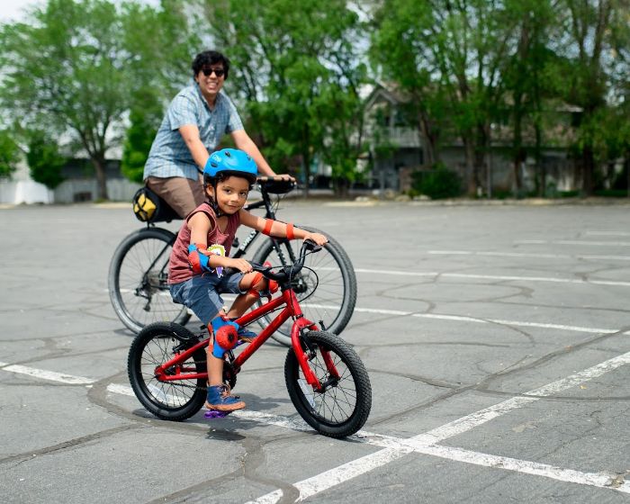 Right side view of a child on a BMX bike, in a parking lot, with a cyclist riding a Surly Bridge Club bike behind them