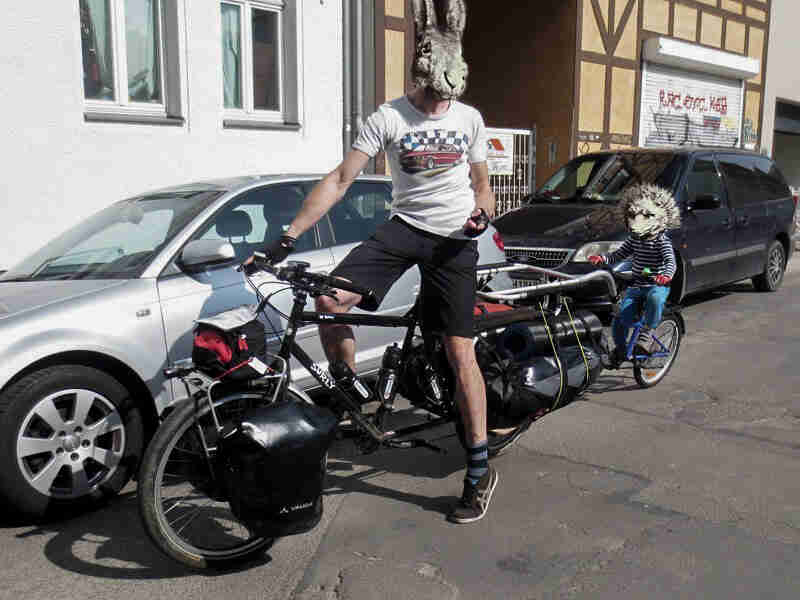 A cyclist wearing a rabbit mask, on a Surly Big Dummy bike, on a street next to a parked car - left front view